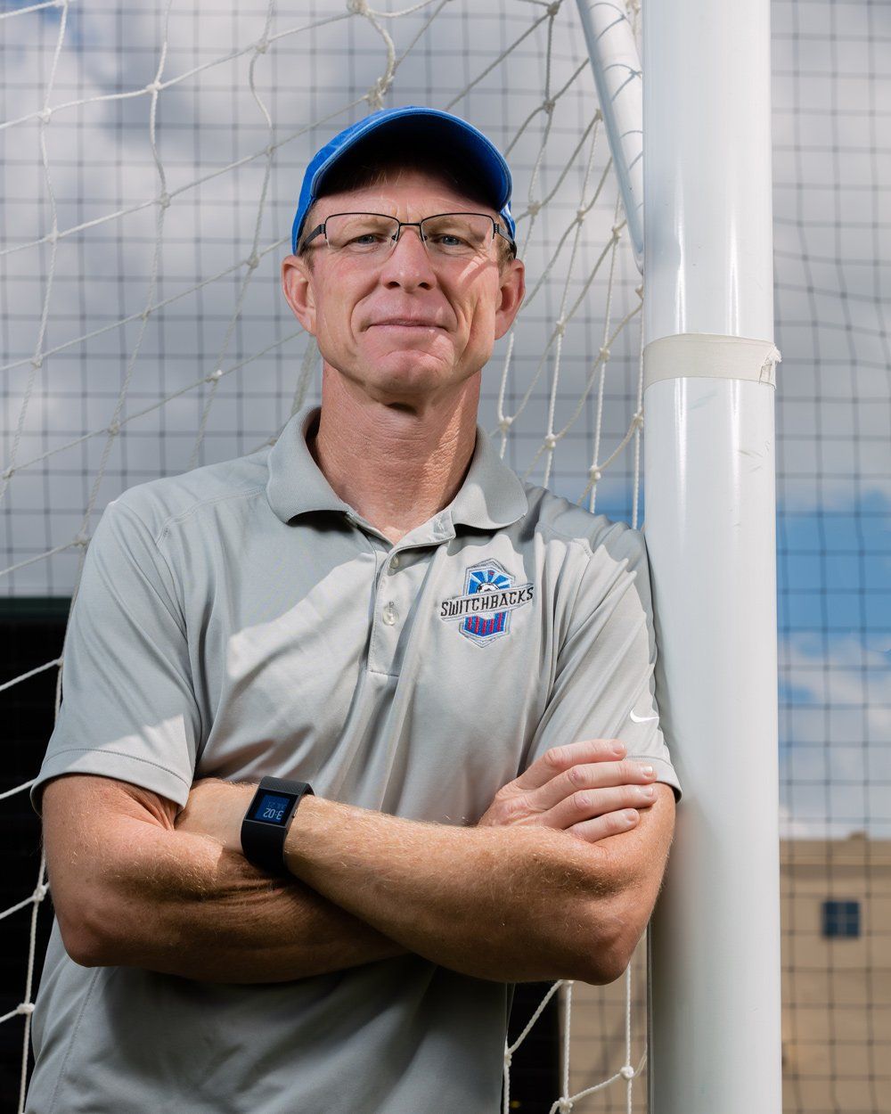Man wearing a blue cap, arms crossed, leaning against a white soccer goalpost, outdoors.