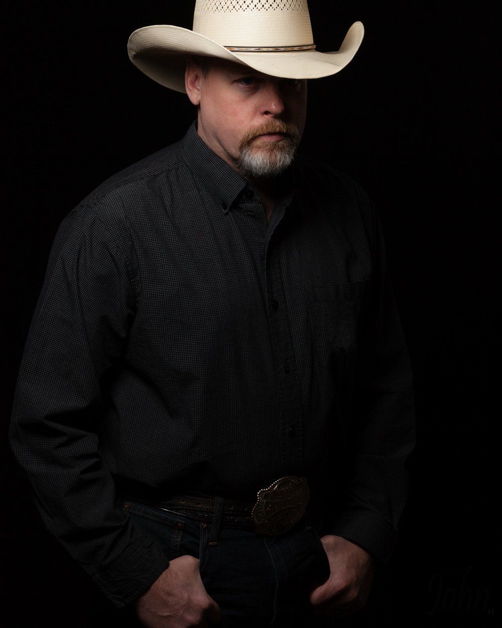 Man wearing a cowboy hat, black shirt, and belt buckle, hands in pockets, looking toward the viewer.
