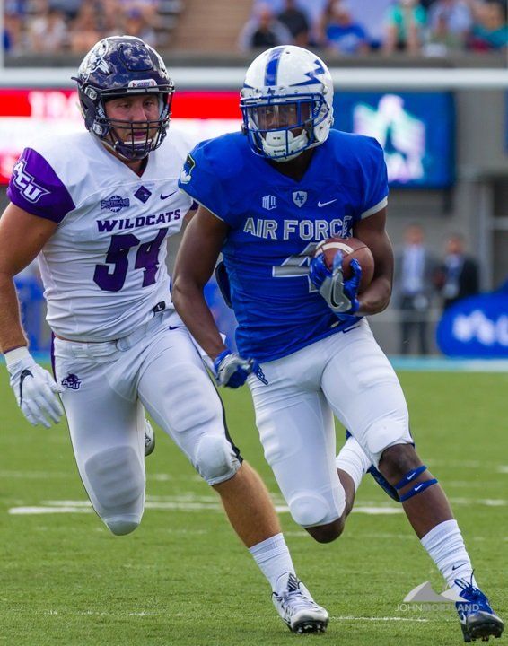 Football player in blue uniform runs with the ball, pursued by a player in purple uniform.