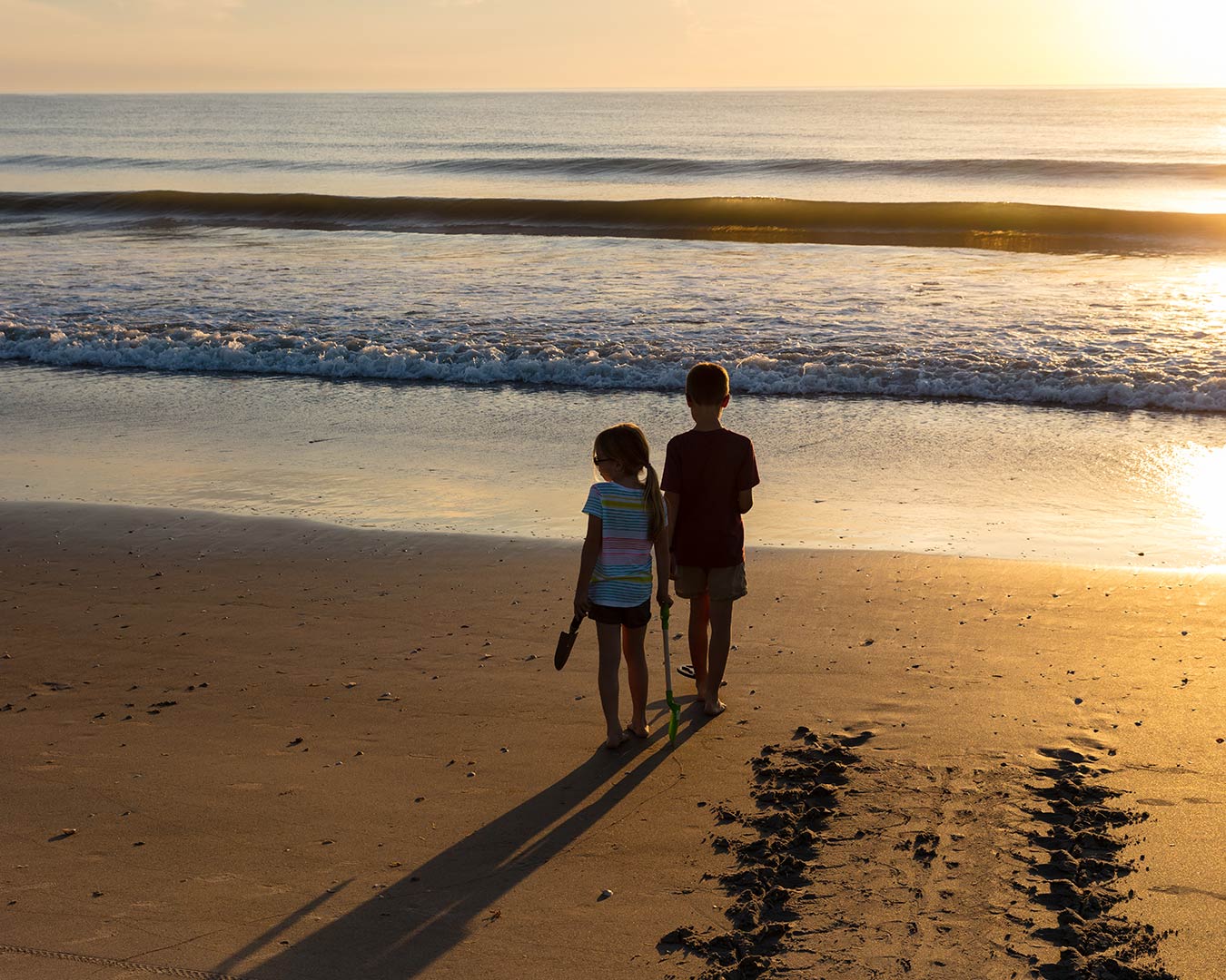 Two children walk on a sandy beach towards the ocean at sunset, casting long shadows.