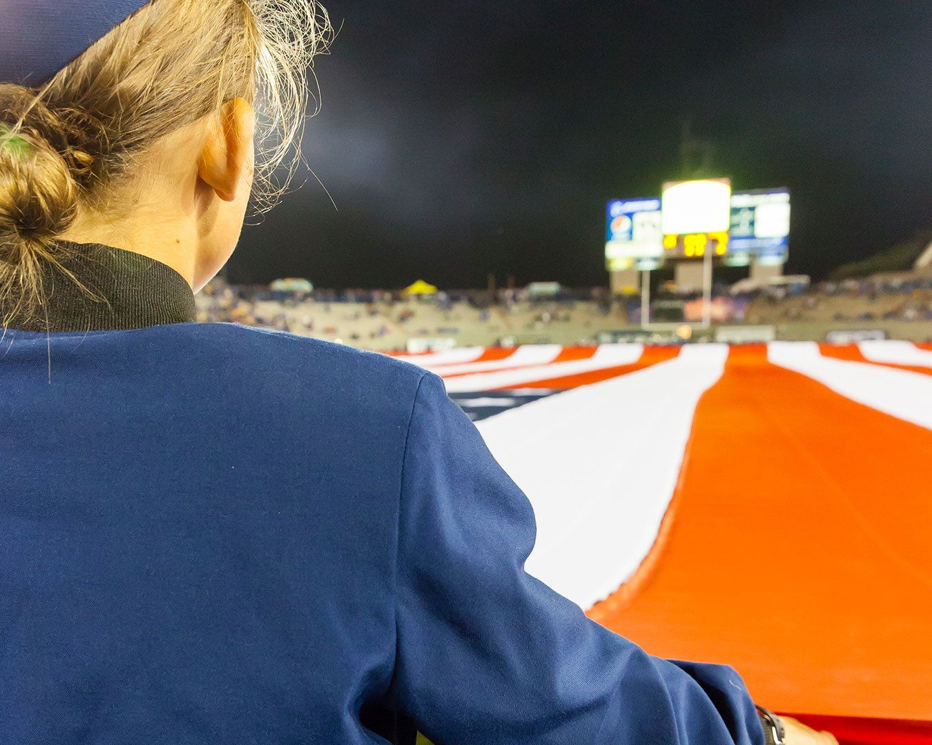 Person holding a giant American flag on a stadium field, watching a scoreboard at night.