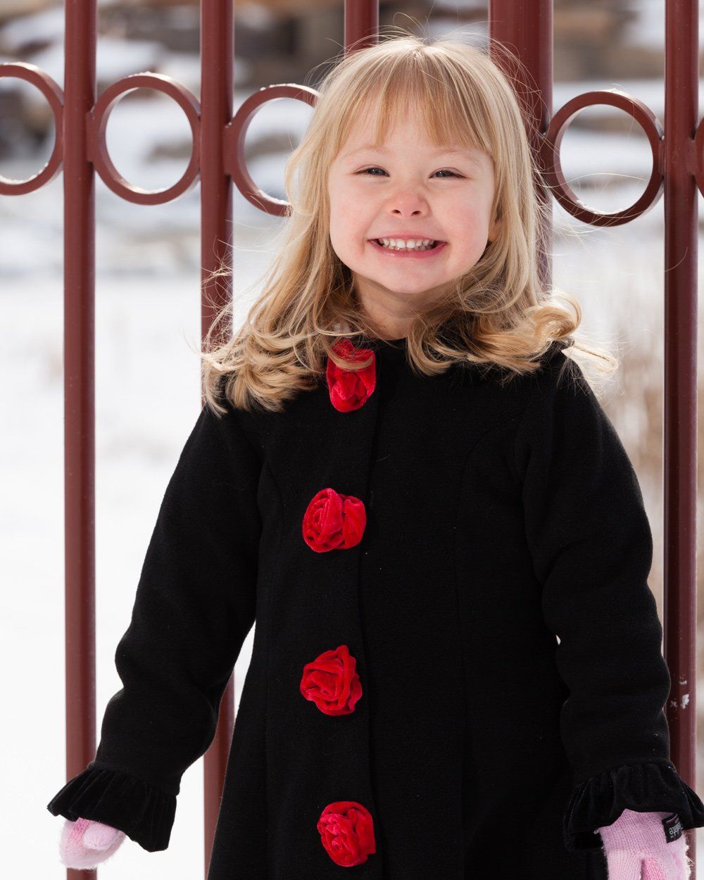 Smiling child in black coat with red flower buttons stands in front of a red fence.