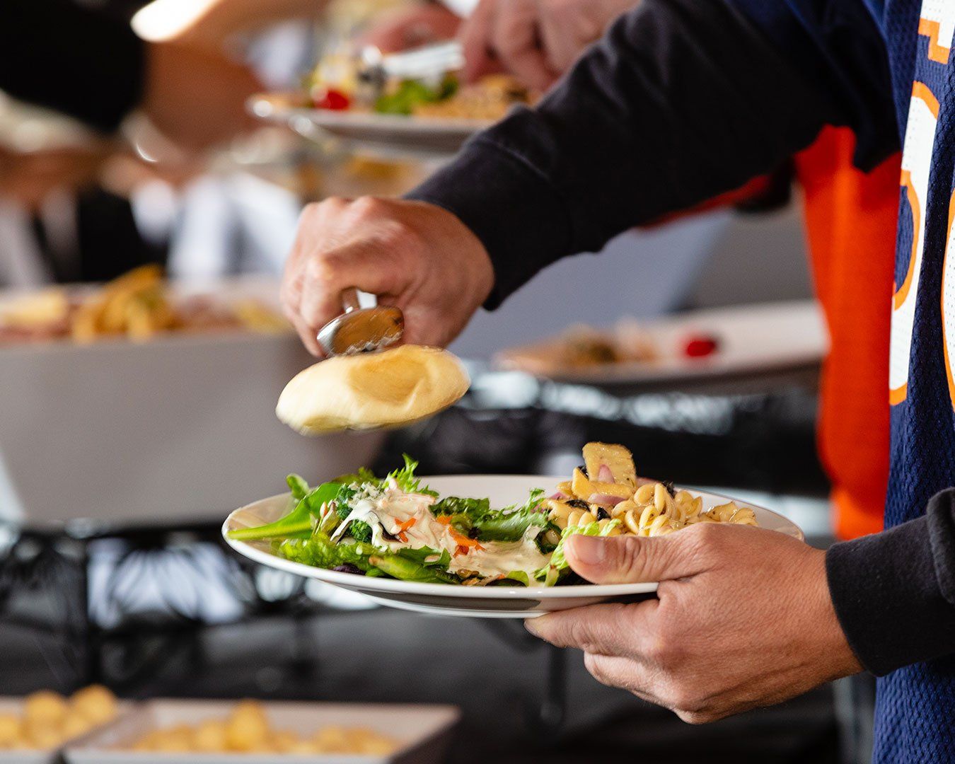 Person at buffet holding a plate of food, adding a roll with tongs; orange and navy jersey visible.