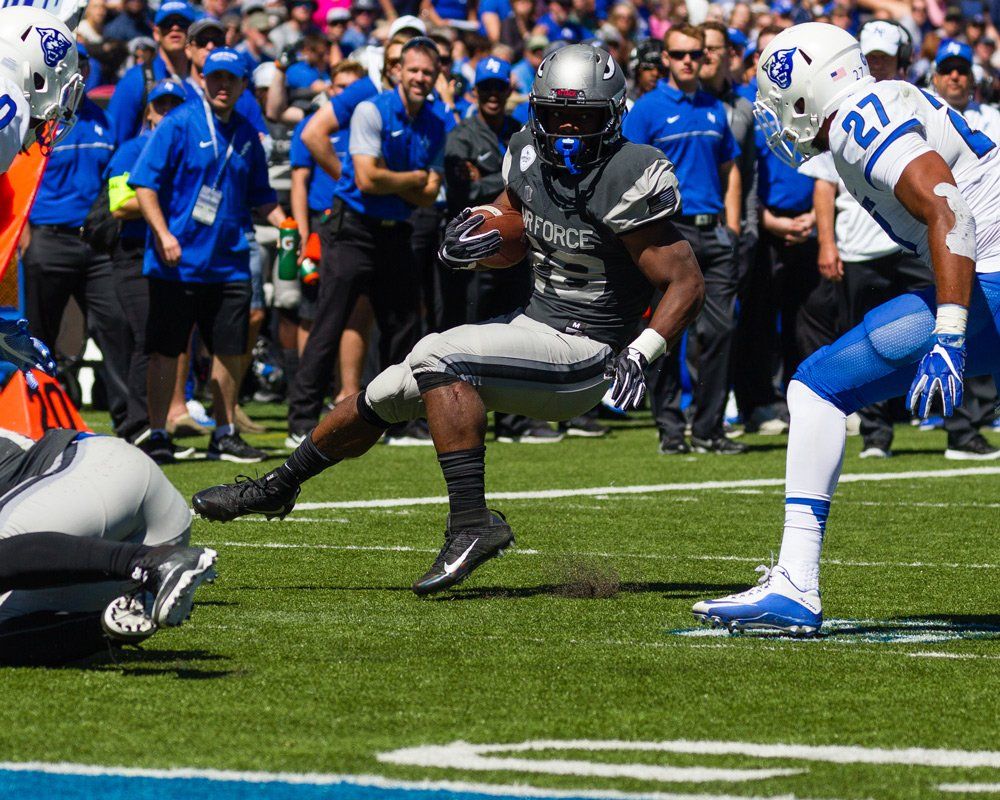 Football player in gray uniform runs with ball, other players nearby. Field, sunny day.
