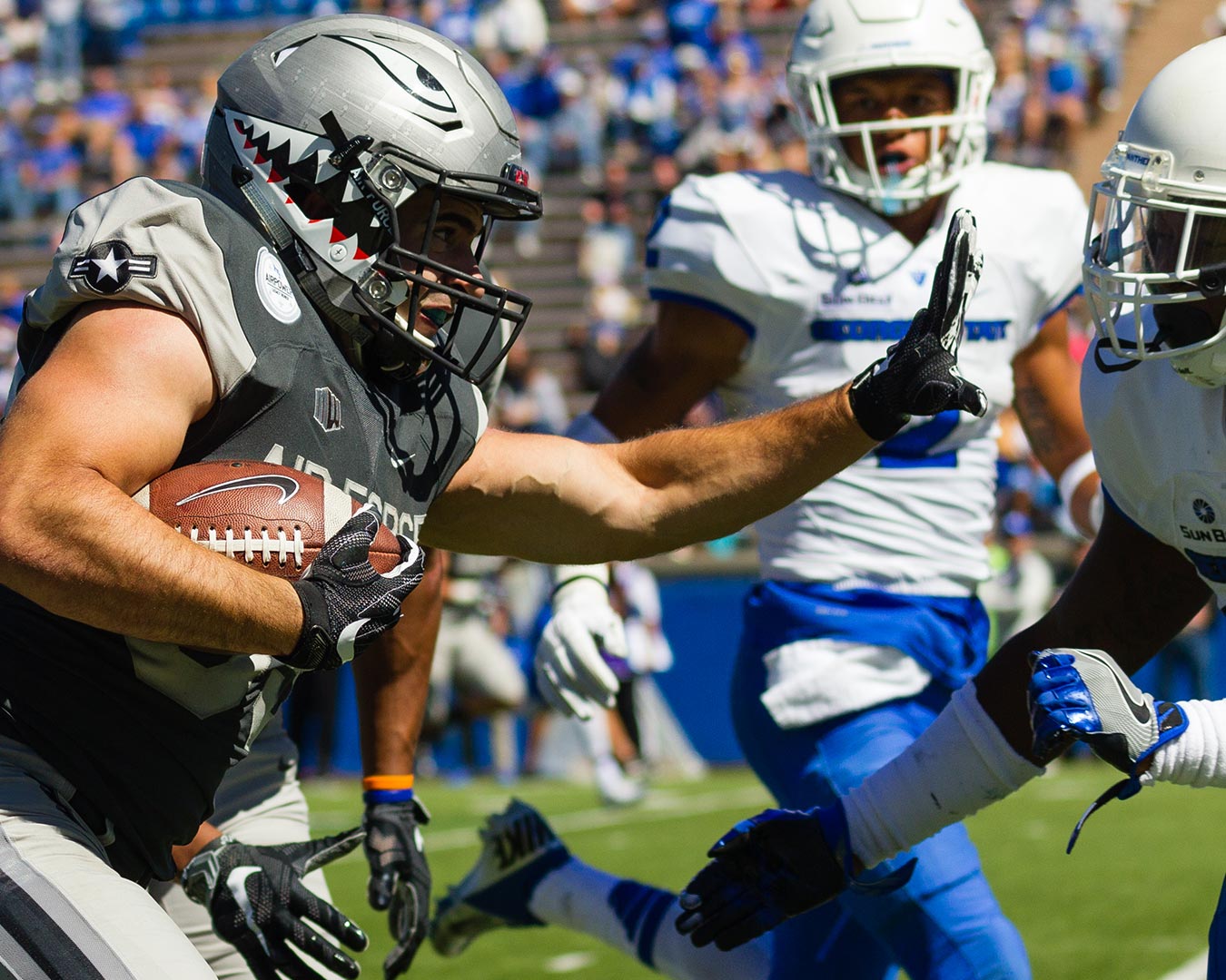 Football player in grey shark-themed helmet runs with ball, fending off blue-clad opponent on field.