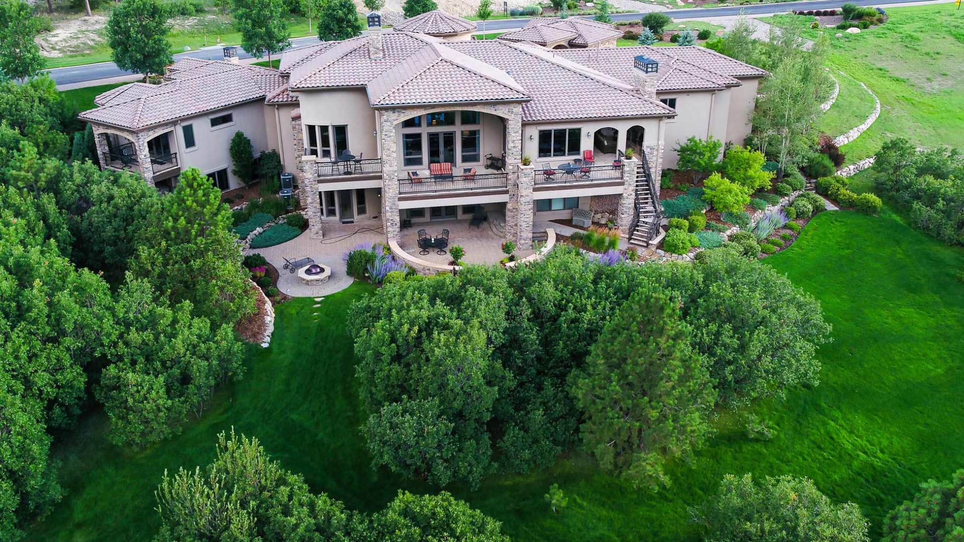 Large stone house with terracotta roof, multiple balconies, surrounded by green trees and grass.
