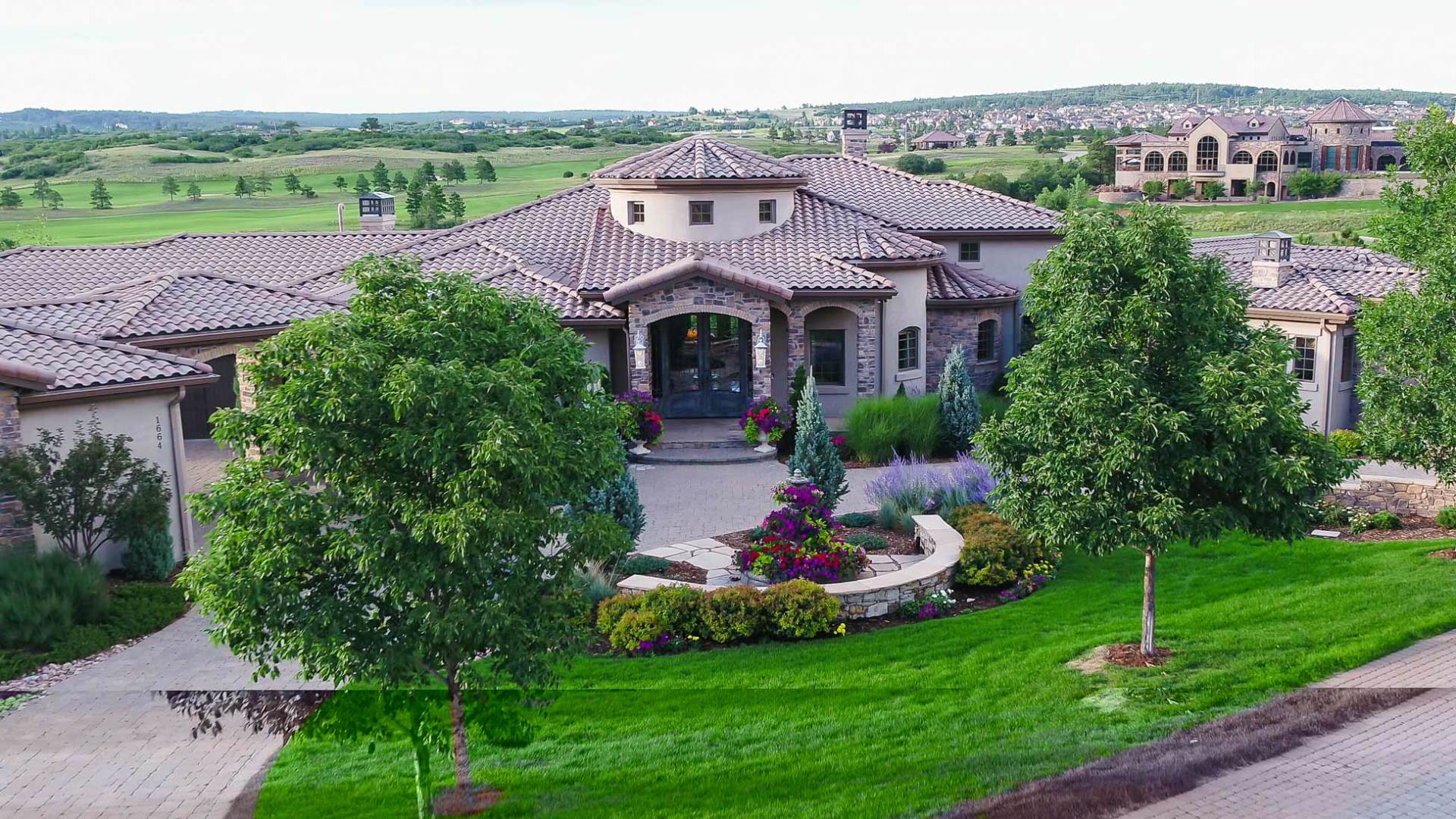 Large house with tile roof, stone facade, and circular driveway, surrounded by green lawns and trees.