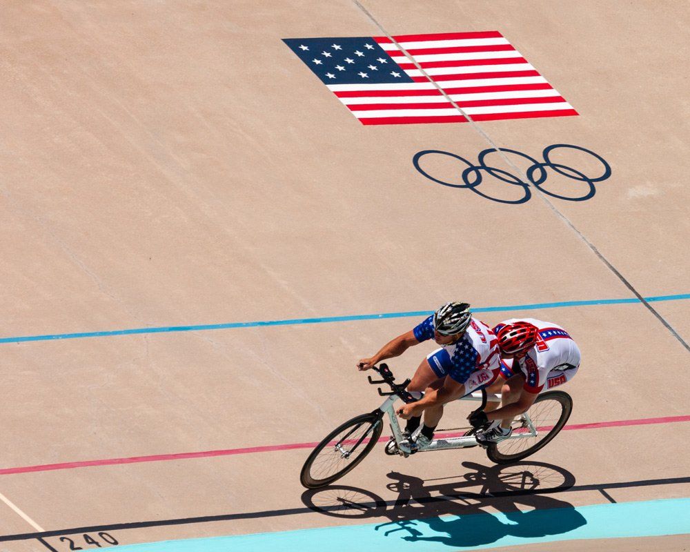 Two cyclists racing on a velodrome with Olympic rings and a U.S. flag painted on the track.