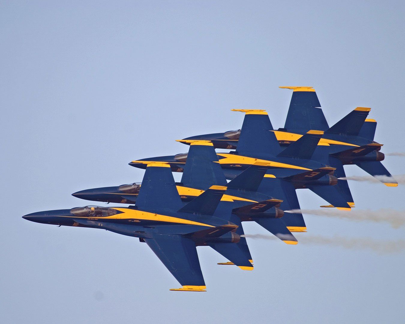 Four blue and yellow jets in tight formation against a pale blue sky, leaving white vapor trails.