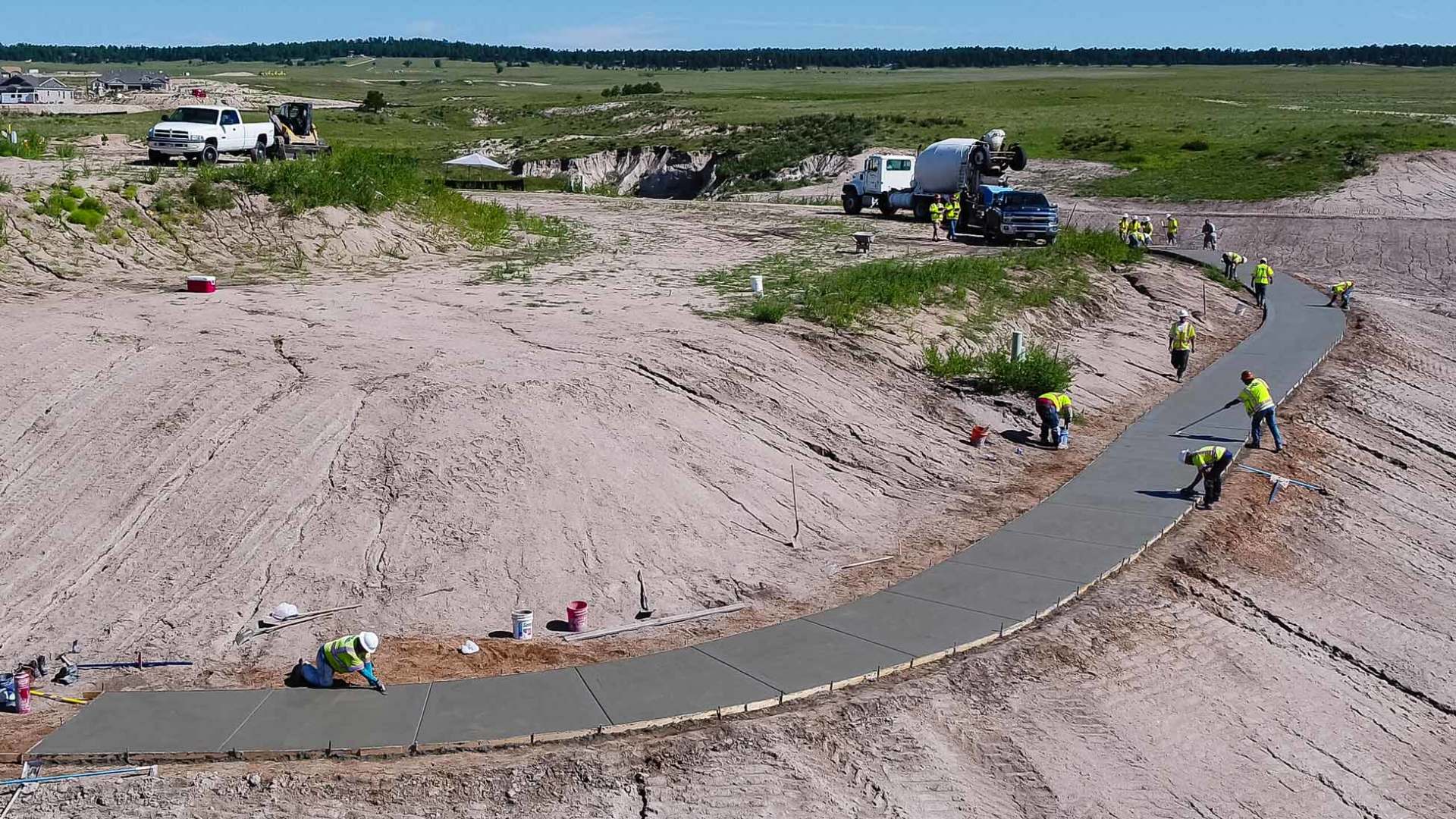 Construction workers pouring and smoothing concrete on a winding pathway in a sandy landscape.
