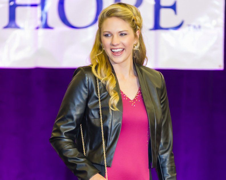 Woman in black jacket and pink dress smiling, on stage with purple backdrop.