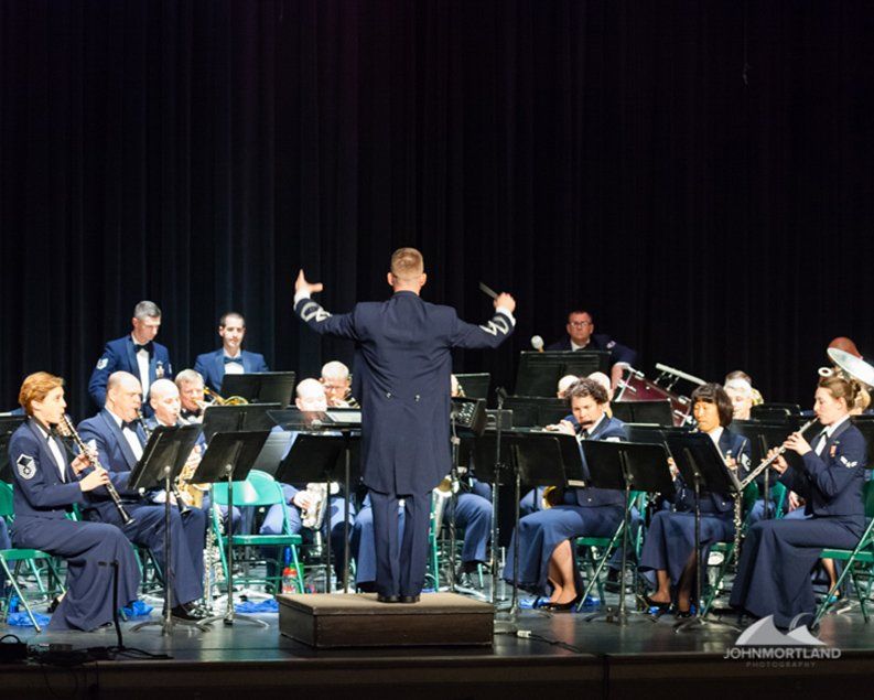 Air Force band performing on stage; conductor leading with arms raised.