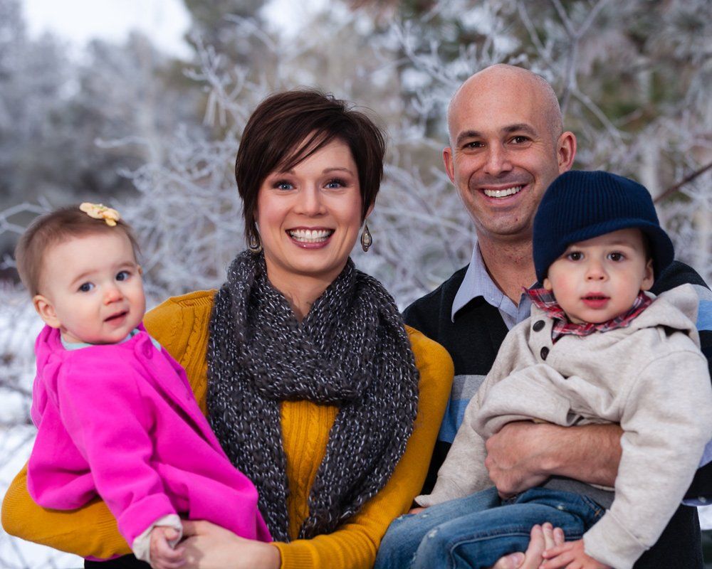 Family of four smiling outdoors: woman in yellow holds baby in pink, man holds child in a hat. Winter background.