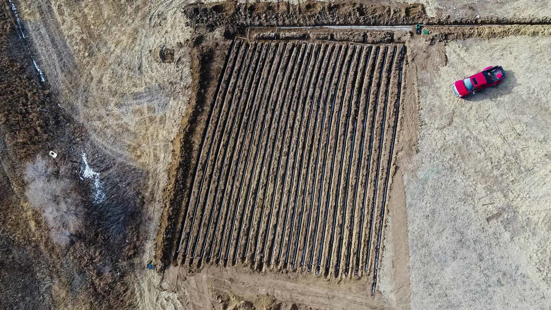Aerial view of a rectangular plowed field with rows, a red car parked nearby, and dry surrounding vegetation.