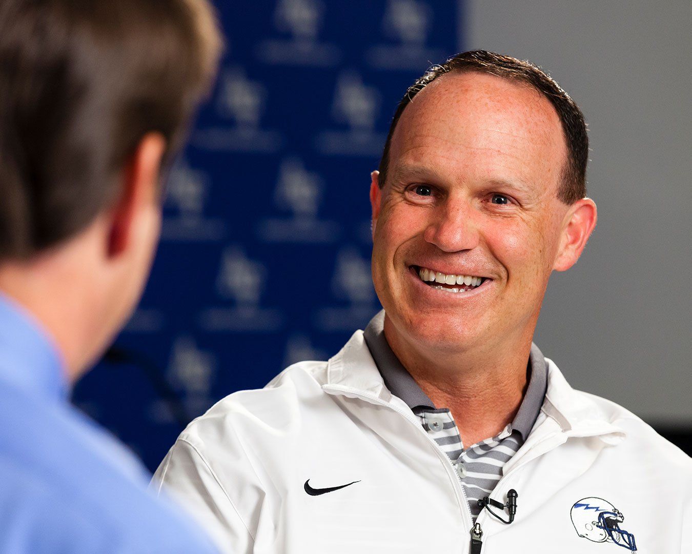 Man smiling during an interview, wearing a white jacket with a grey collar. Background is blue.