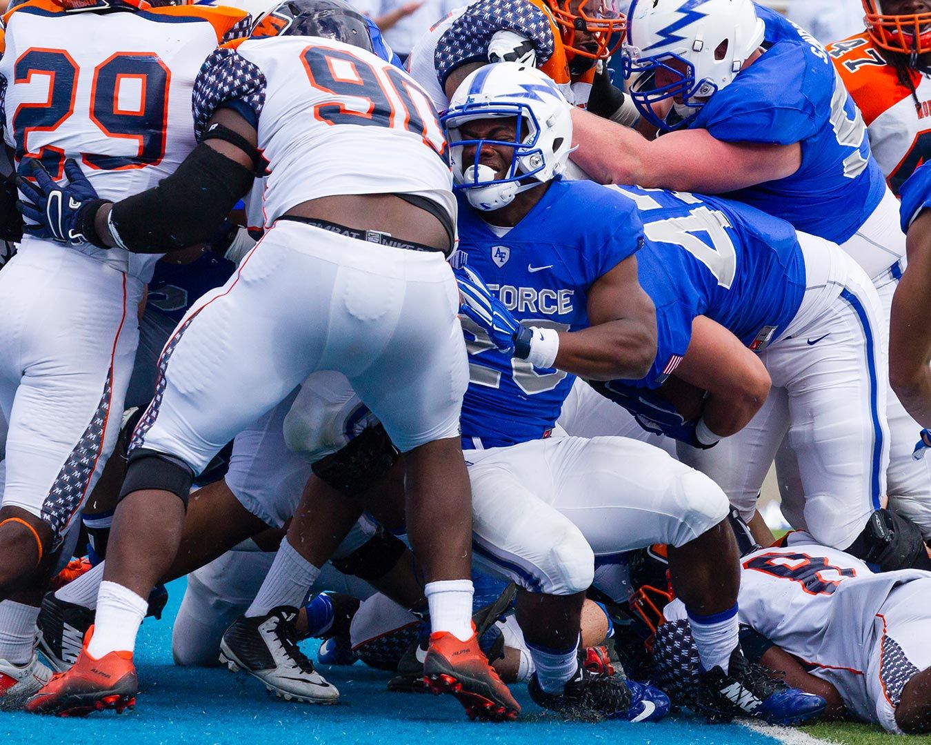 Football players in scrimmage, blue and orange uniforms, intense action.