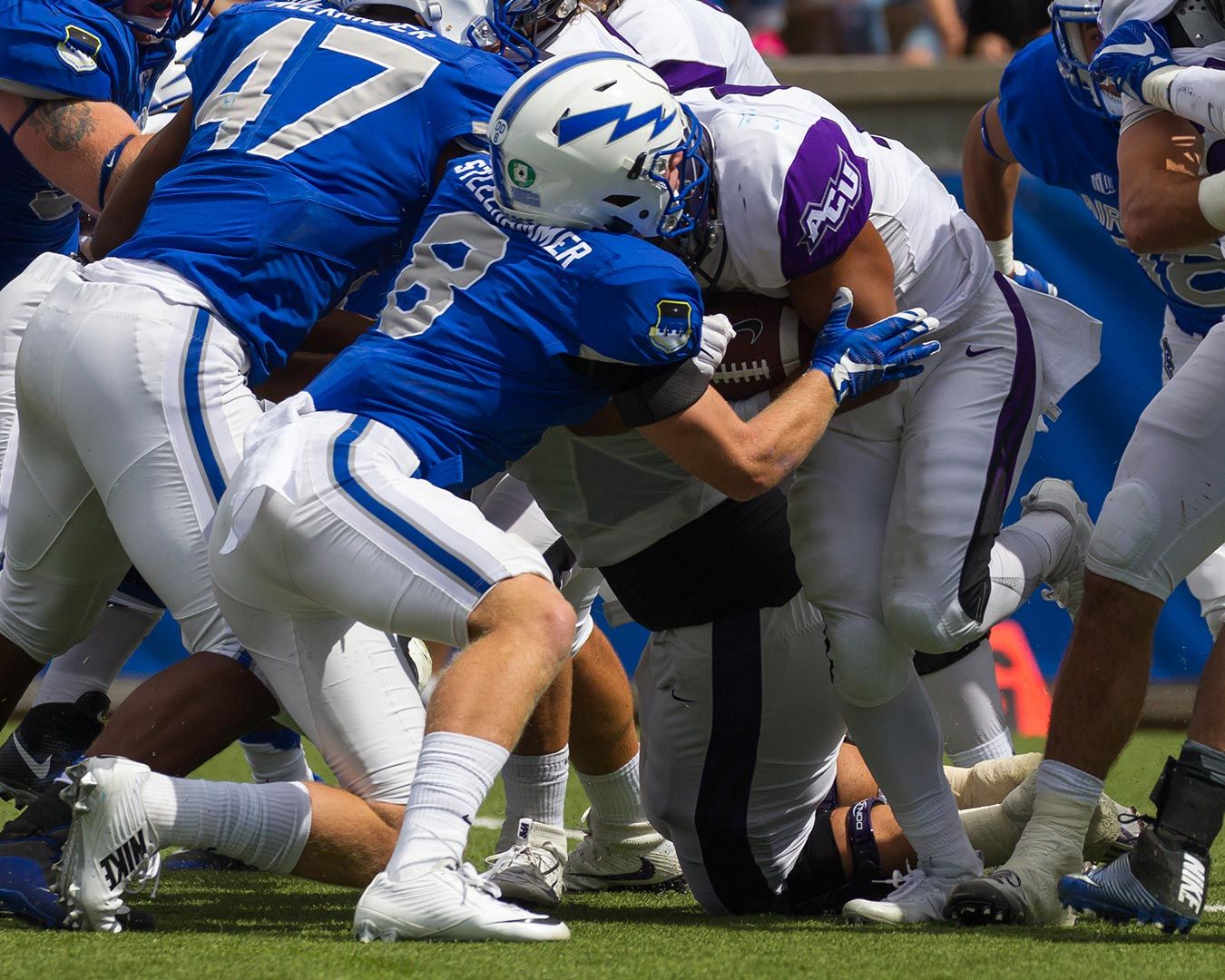Football players in blue and white uniforms tackle a player with the ball, on a green field.