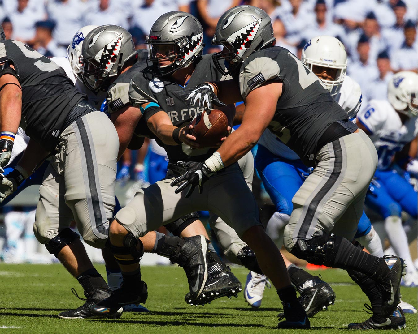 Football players in grey uniforms, one with ball, tackled by players in blue; sunny stadium.