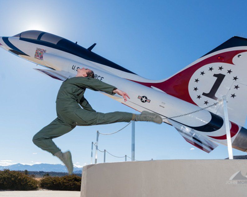 A person in a jumpsuit leaps in front of a white, red, and black airplane on a sunny day.