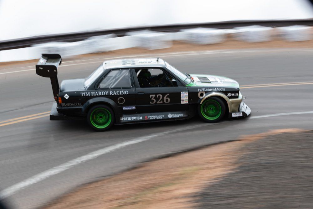 Black race car with green wheels, number 326, speeding around a mountain curve.