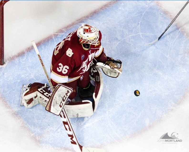 Hockey goalie in burgundy jersey, sprawled in net, watching puck.