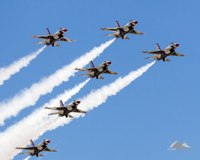 Six gray fighter jets with red accents fly in formation, leaving white contrails against a blue sky.