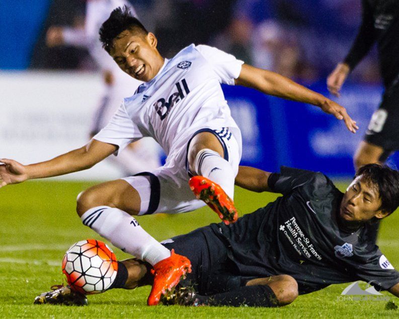Soccer players in action: white-shirted player sliding tackles black-shirted player, ball on the ground.