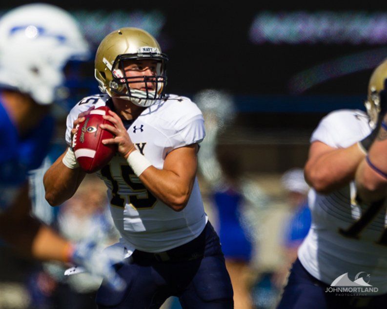 Navy football player in white and gold uniform preparing to throw the ball during a game.