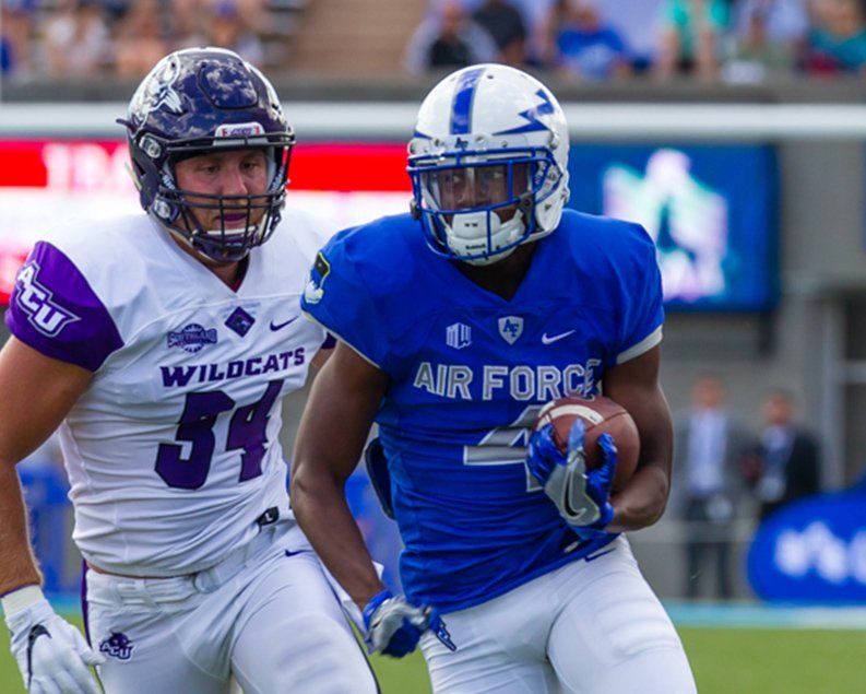 Football player in blue uniform runs with ball, pursued by player in white Wildcats uniform. Stadium background.