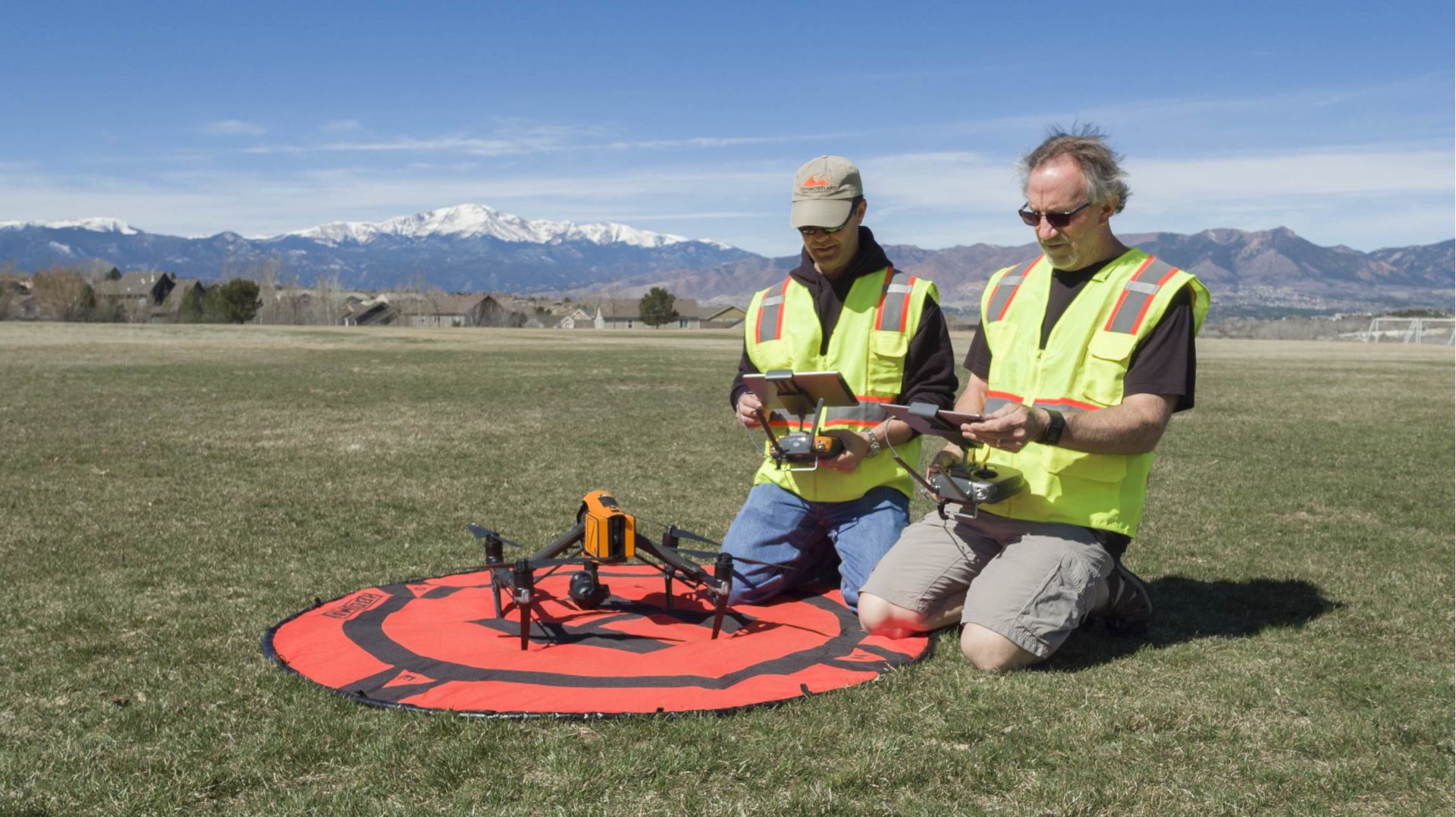 Two people kneeling, operating drones on a red landing pad in a grassy field with mountains in the background.