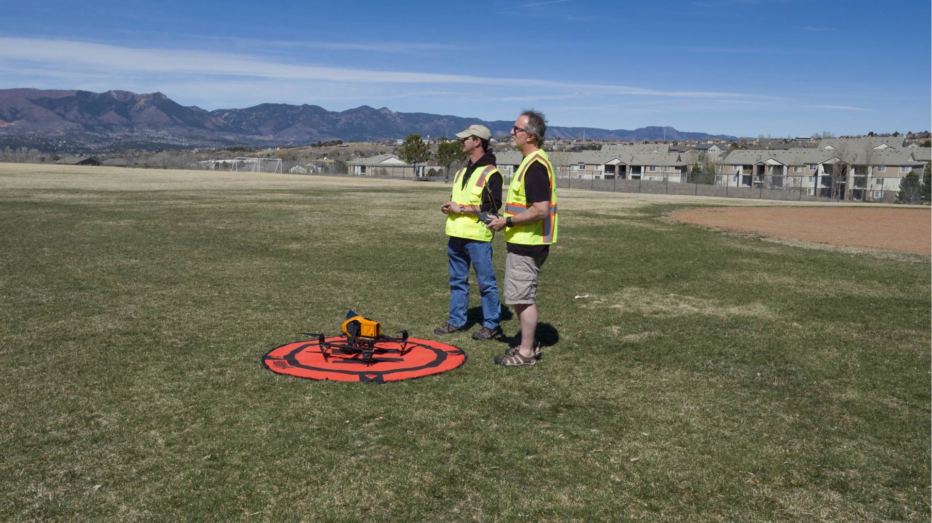Two people with remote controls standing near a drone on a landing pad in a grassy field. Mountains in background.