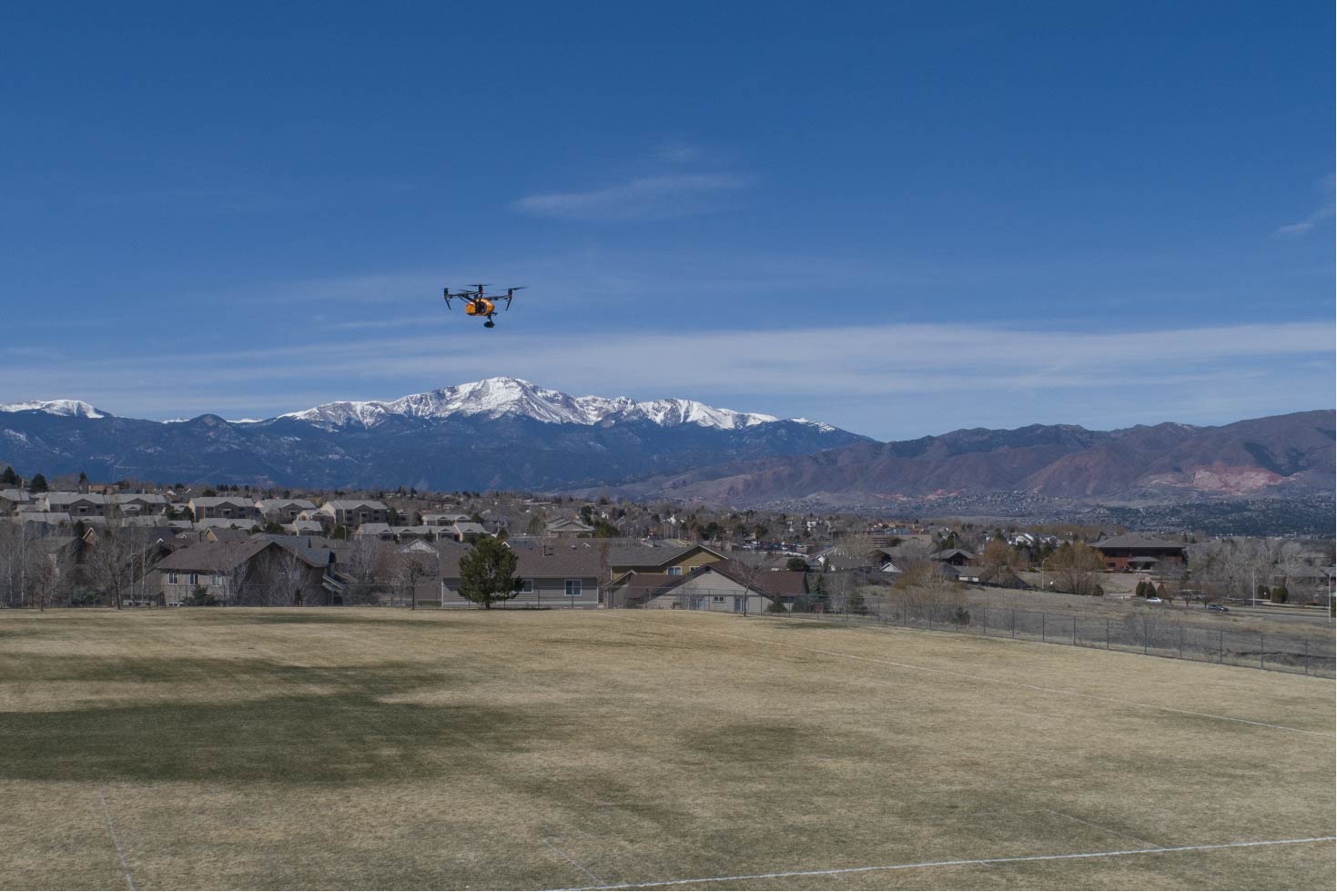 Drone flying over a town with snow-capped mountains in the background, under a blue sky.