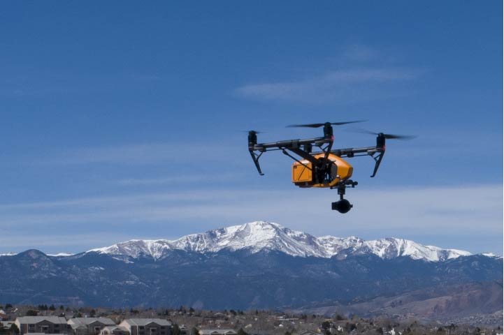Drone carrying cargo in flight, with snow-capped mountain backdrop and blue sky.
