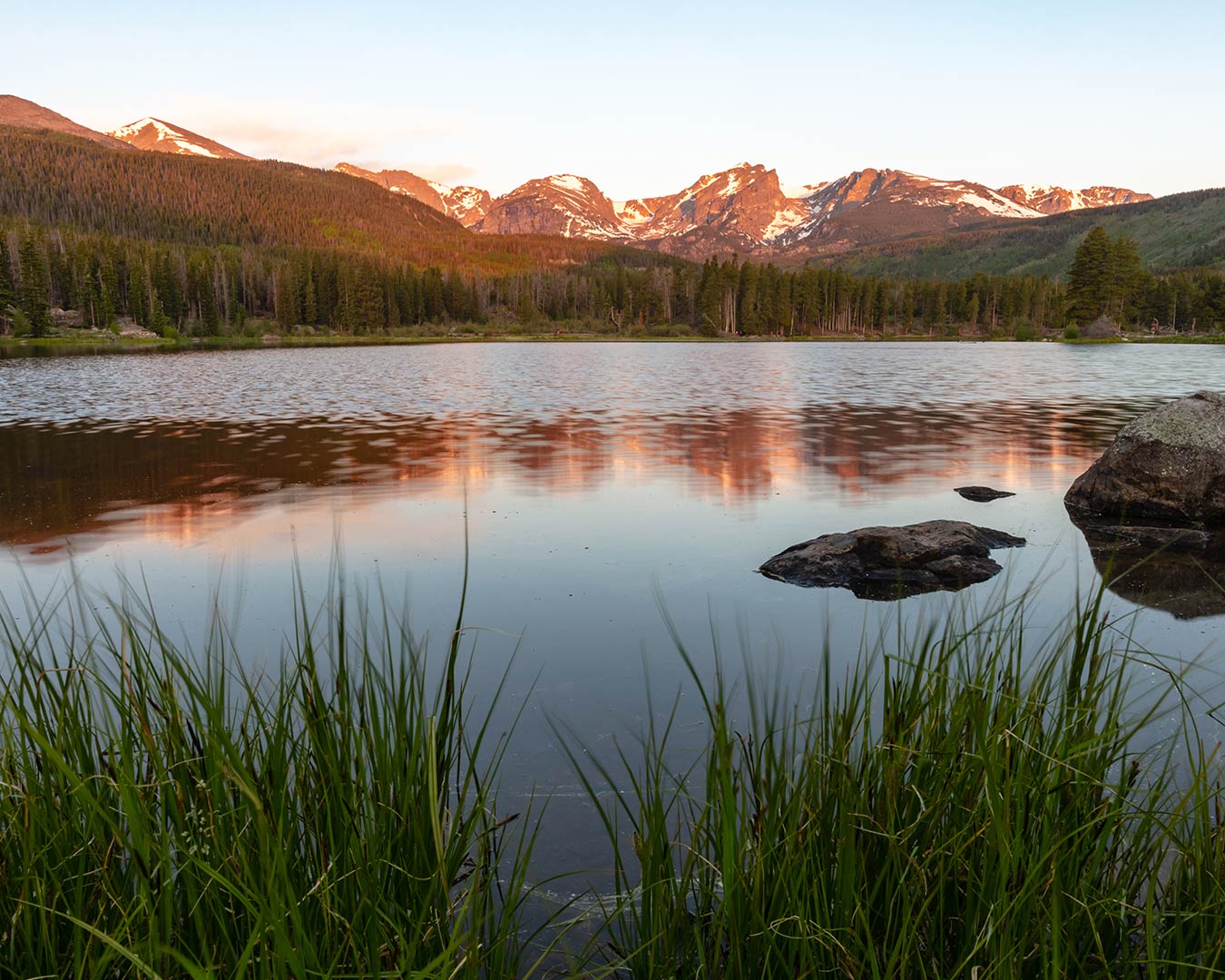Lake reflecting mountains and trees at sunset, with grass in the foreground.