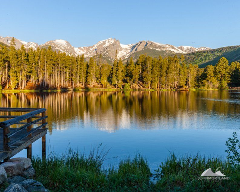 Lakeside view with a wooden dock, tall trees, and mountains reflecting on the water under a blue sky.