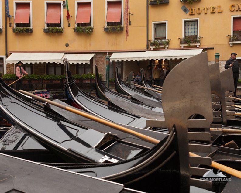 Gondolas lined up in Venice, Italy; a woman rows one. Yellow building with red awnings in the background.