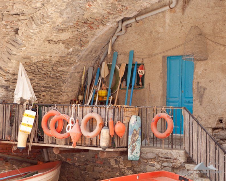 Coastal scene: wooden boats below a stone building with blue door, life rings, and fishing gear.