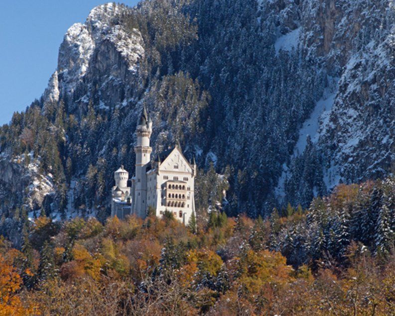 Neuschwanstein Castle nestled in autumn trees against a snow-covered mountain.