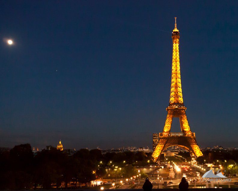 Eiffel Tower illuminated at night against a dark blue sky with a visible moon.