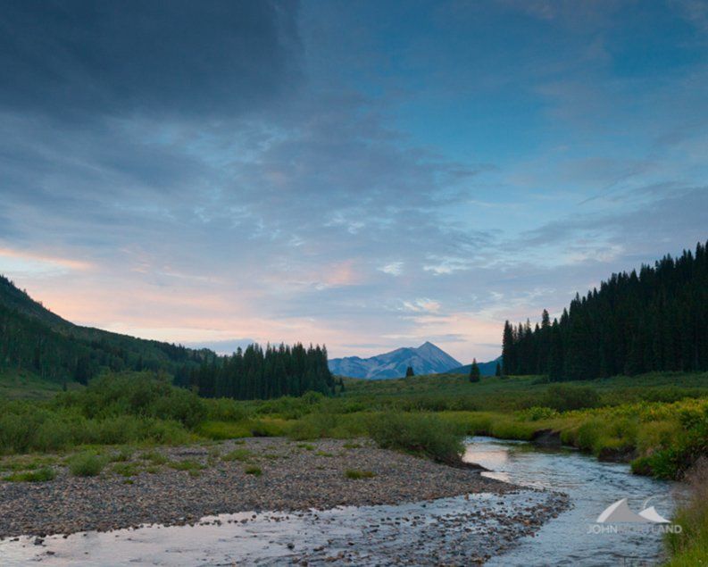 River flowing through a green meadow with a mountain in the distance under a cloudy sky.