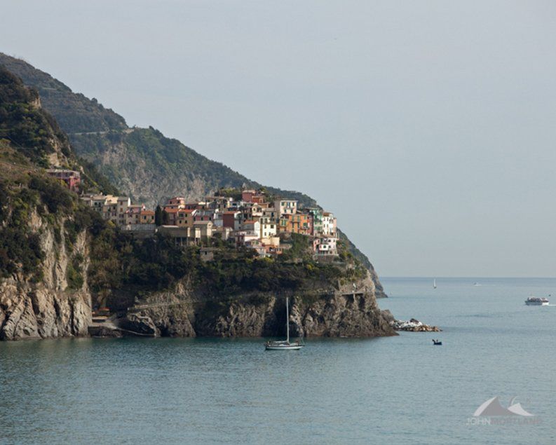 Coastal village on a cliff overlooking the sea, with colorful buildings and a sailboat.