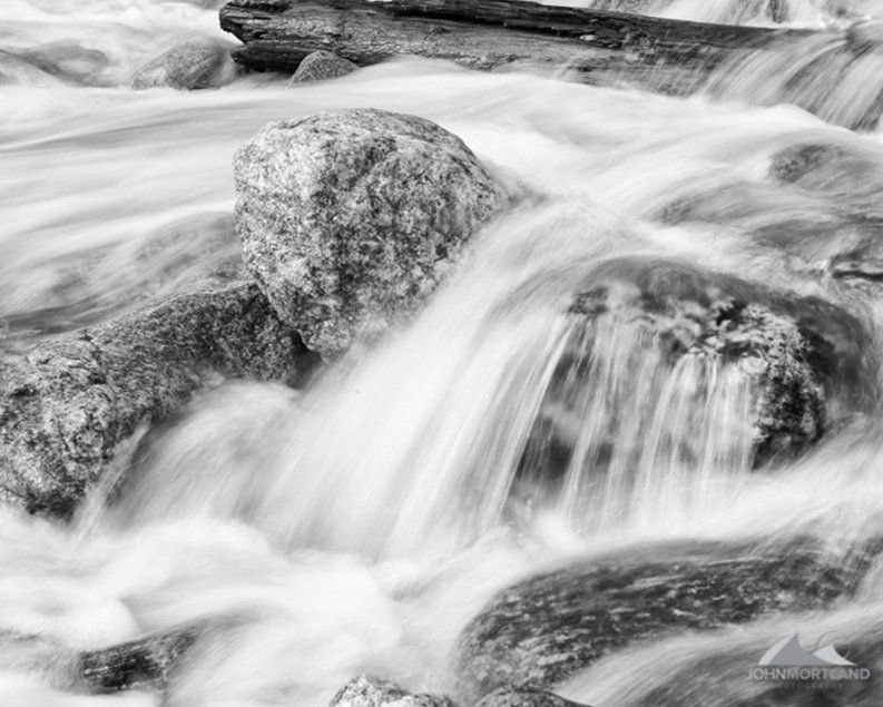 Black and white photo of water cascading over rocks in a stream. Long exposure blurs the water.