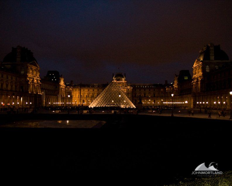 Night view of the Louvre Museum, with the glass pyramid illuminated, and surrounding buildings lit by soft lights.