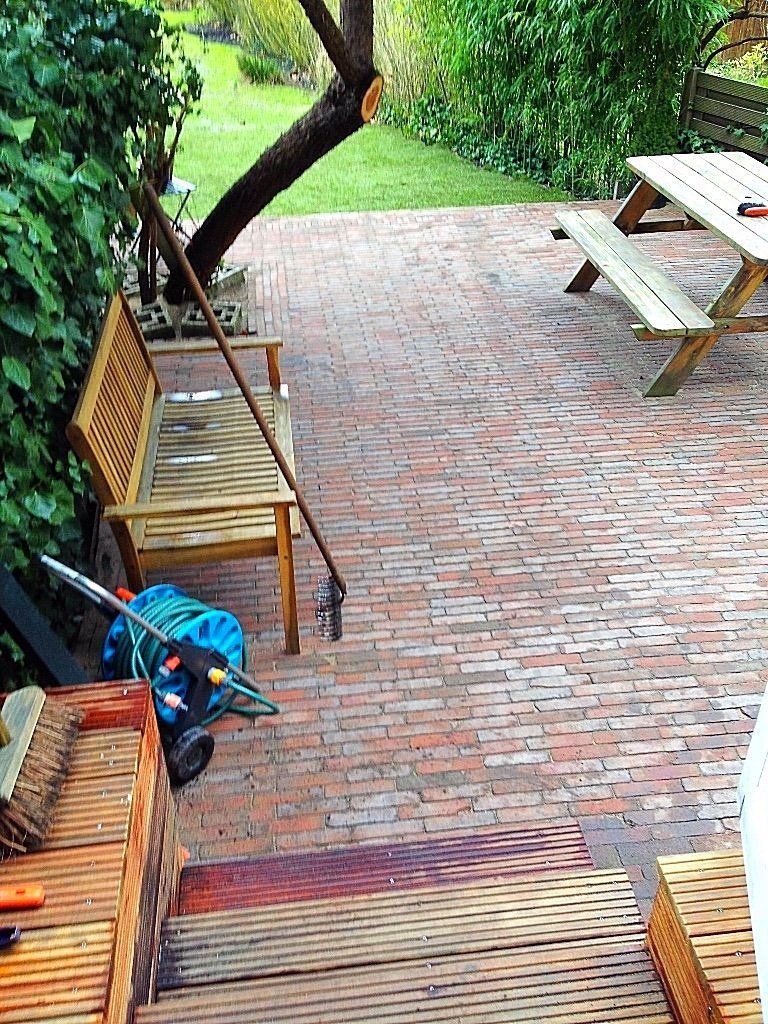 Bakstenen terras met houten bank, picknicktafel en tuingereedschap. Groen gazon en boom op de achtergrond.