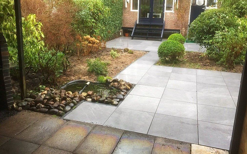 Terras met grijze stenen, een kleine vijver, groen en een huis met een zwarte deur op de achtergrond.