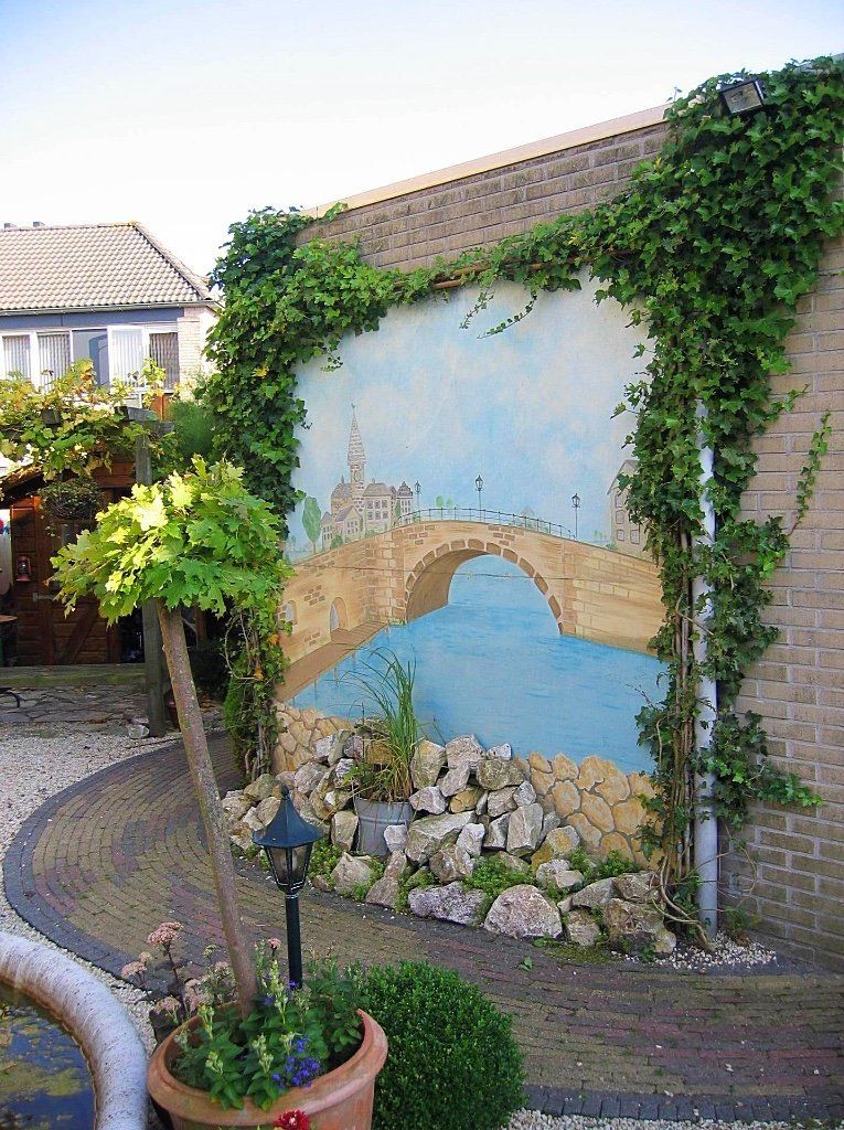 Muurschildering van een brug over water op een bakstenen muur, met groen en potplanten eromheen.