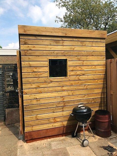 Houten schuur met klein raam, barbecue en ton in de achtertuin.
