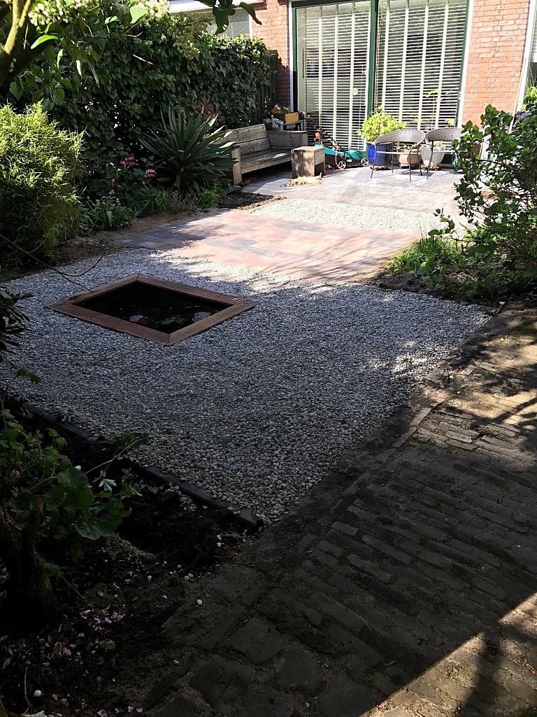 Een grindterras met een vierkante vijver, een bakstenen pad en een bankje in een tuinachtige omgeving.