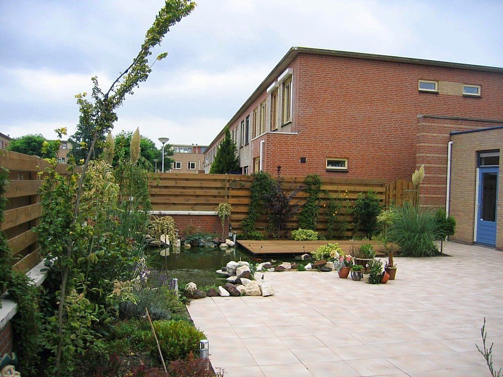 Achtertuin met vijver, houten terras, stenen patio, planten en een bakstenen bijgebouw.