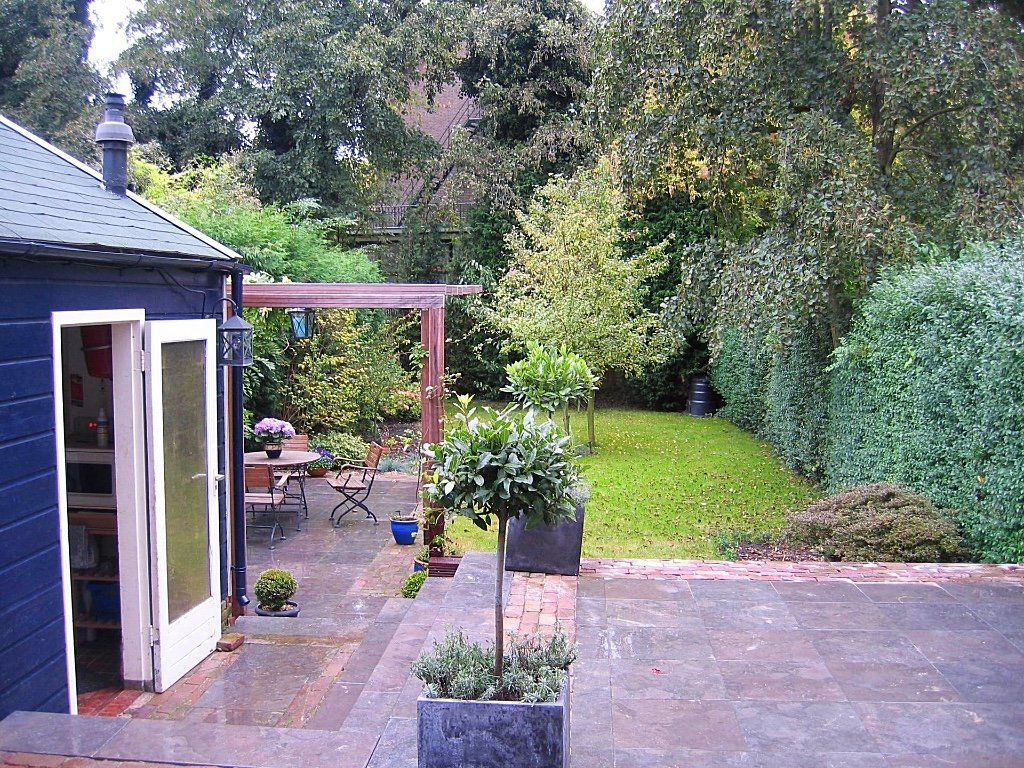 Achtertuin met terras, gazon, bomen en een blauw gebouw met openslaande deuren.